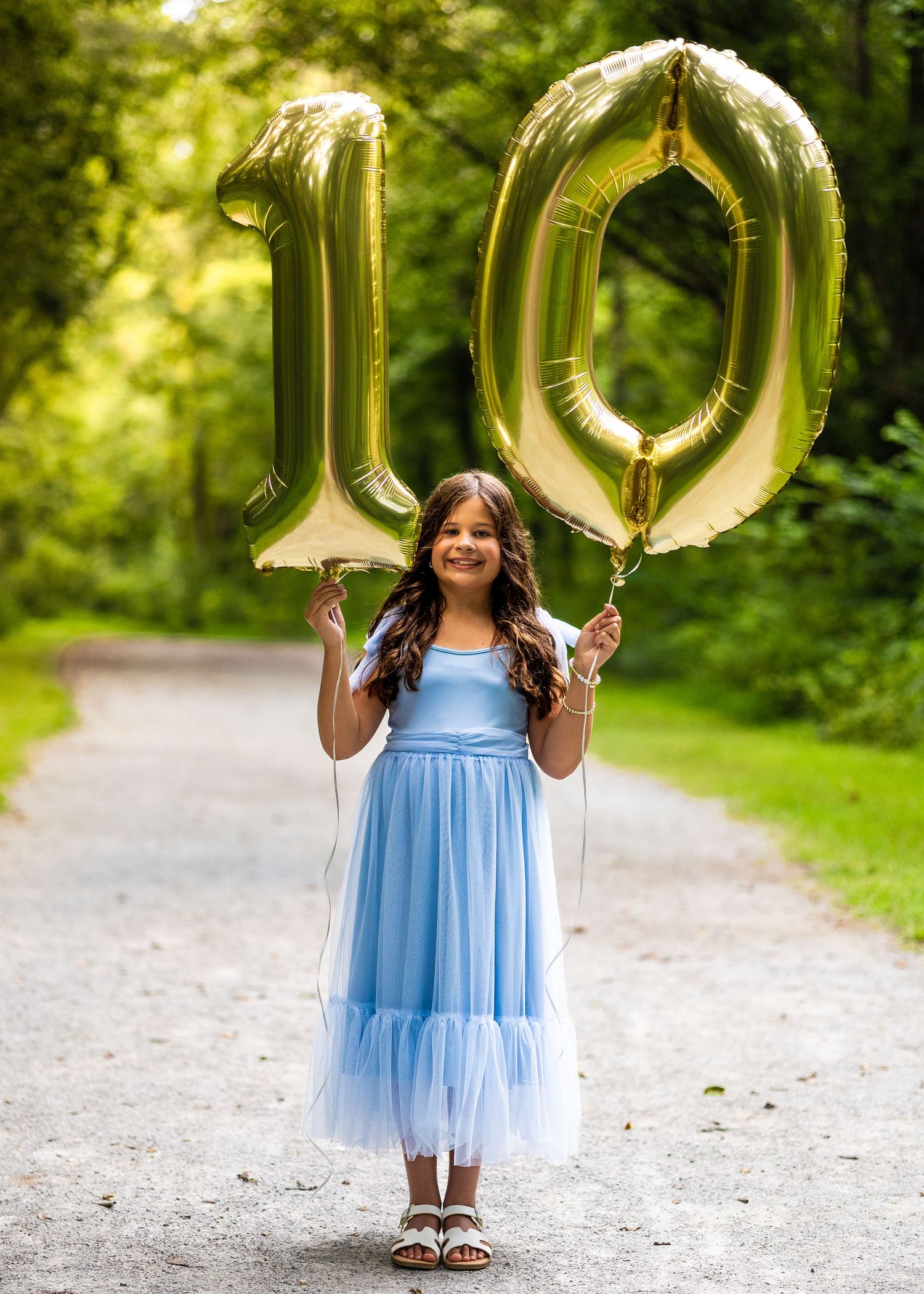 A smiling girl in a blue tulle dress holds gold number 10 balloons on a tree-lined path celebrating her birthday.