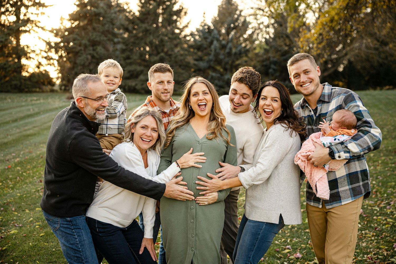 A joyful extended family gathers outdoors at golden hour, celebrating a pregnancy with smiles and laughter in a lush green park.