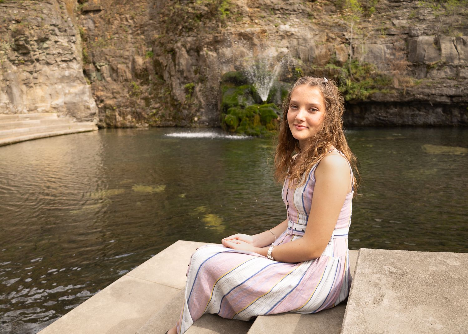 Young woman in striped dress sits smiling by a serene pond with a fountain and rocky cliff backdrop.