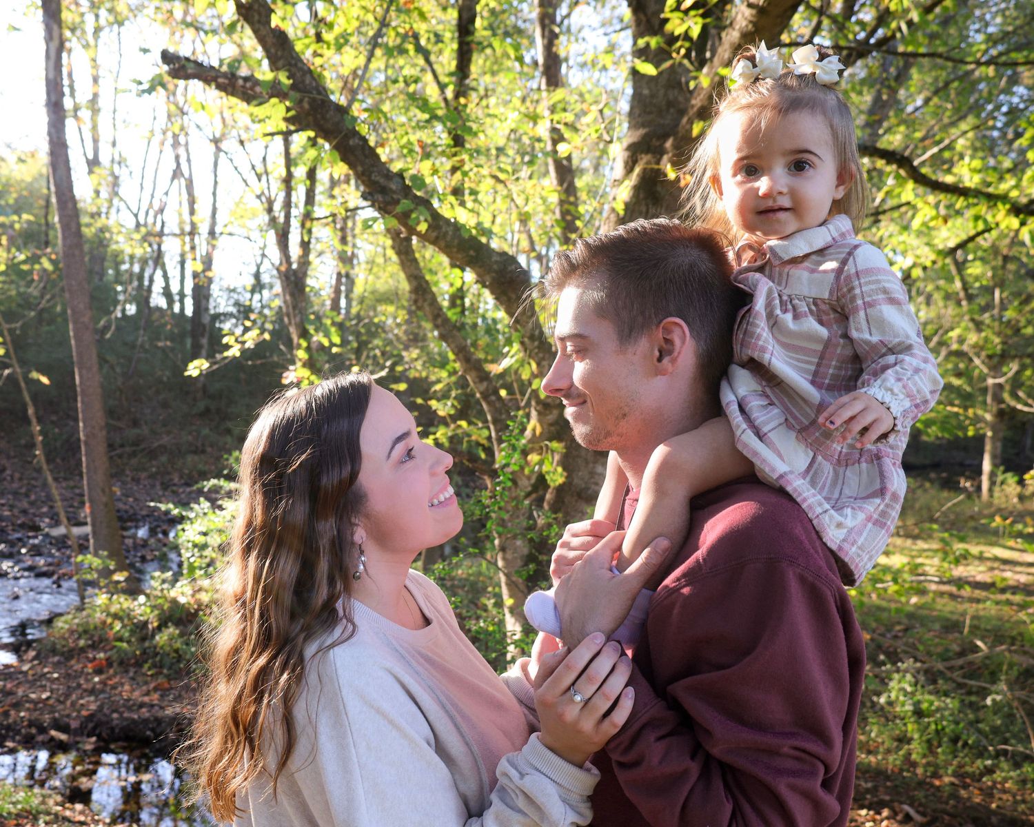 A young family poses outdoors in golden sunlight, with parents smiling and a toddler girl sitting on dad's shoulders.