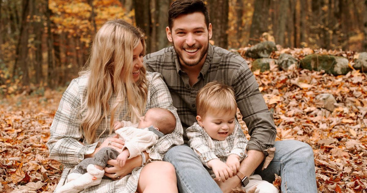 Family of four poses in autumn forest, parents smiling with newborn baby and toddler among fallen orange leaves.