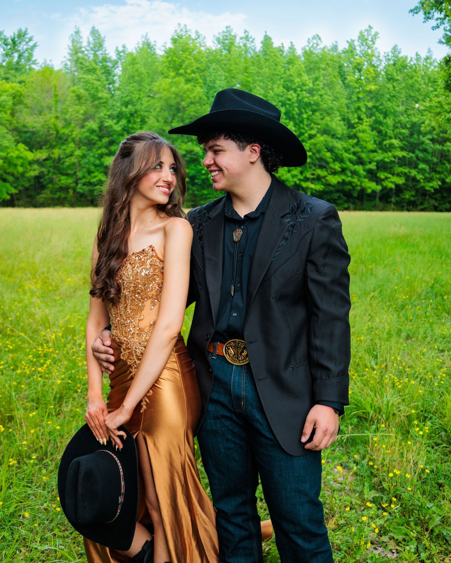A couple in formal western attire poses in a green meadow, she in a gold gown, he in a black suit and cowboy hat.