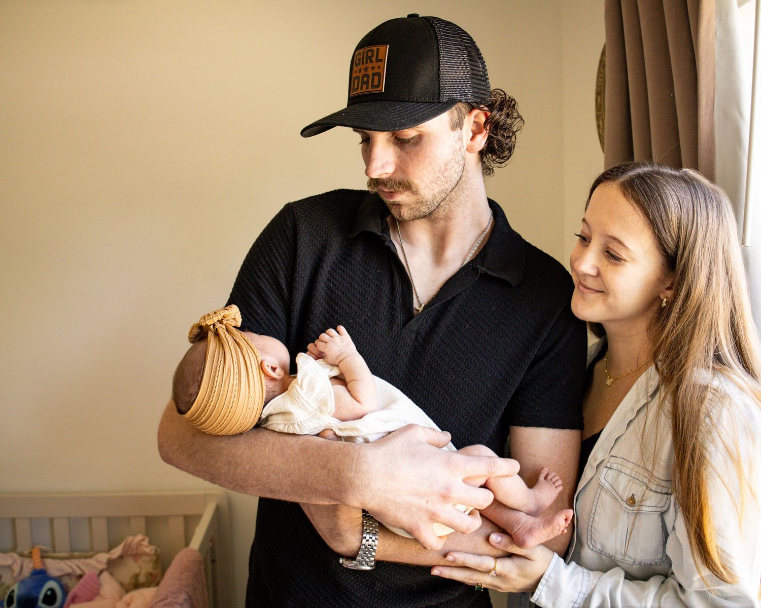 A young couple tenderly holds their newborn baby, the man wearing a black shirt and cap, the woman in a denim jacket.