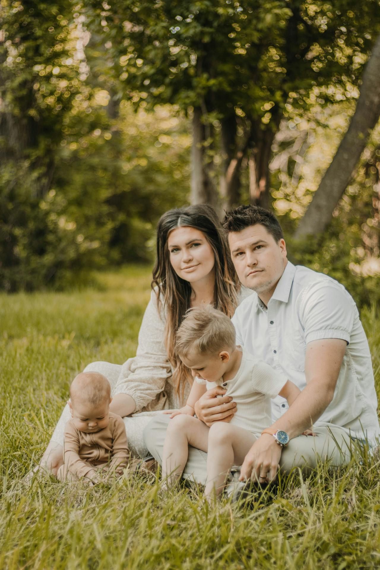 Young family with two toddlers sitting on green grass in a sunny park, parents smiling warmly at the camera.