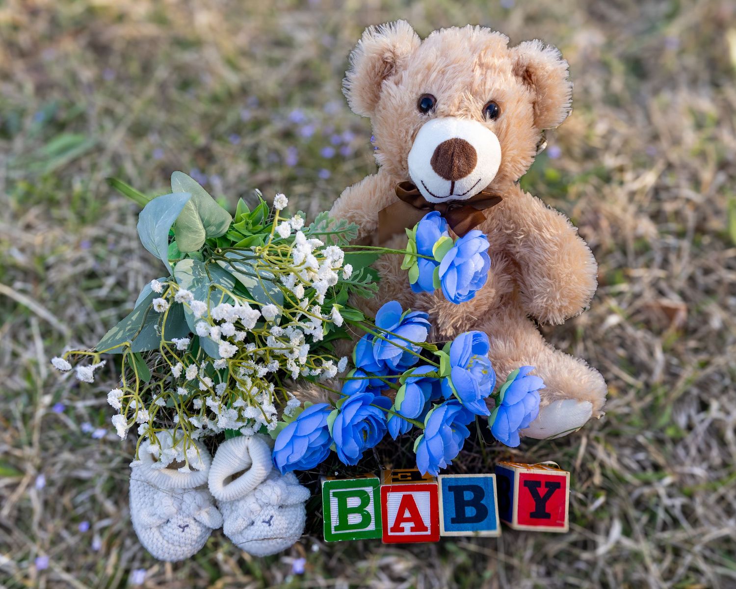 Teddy bear with blue roses, baby blocks spelling BABY, white booties, and floral bouquet on grass for a baby shower.