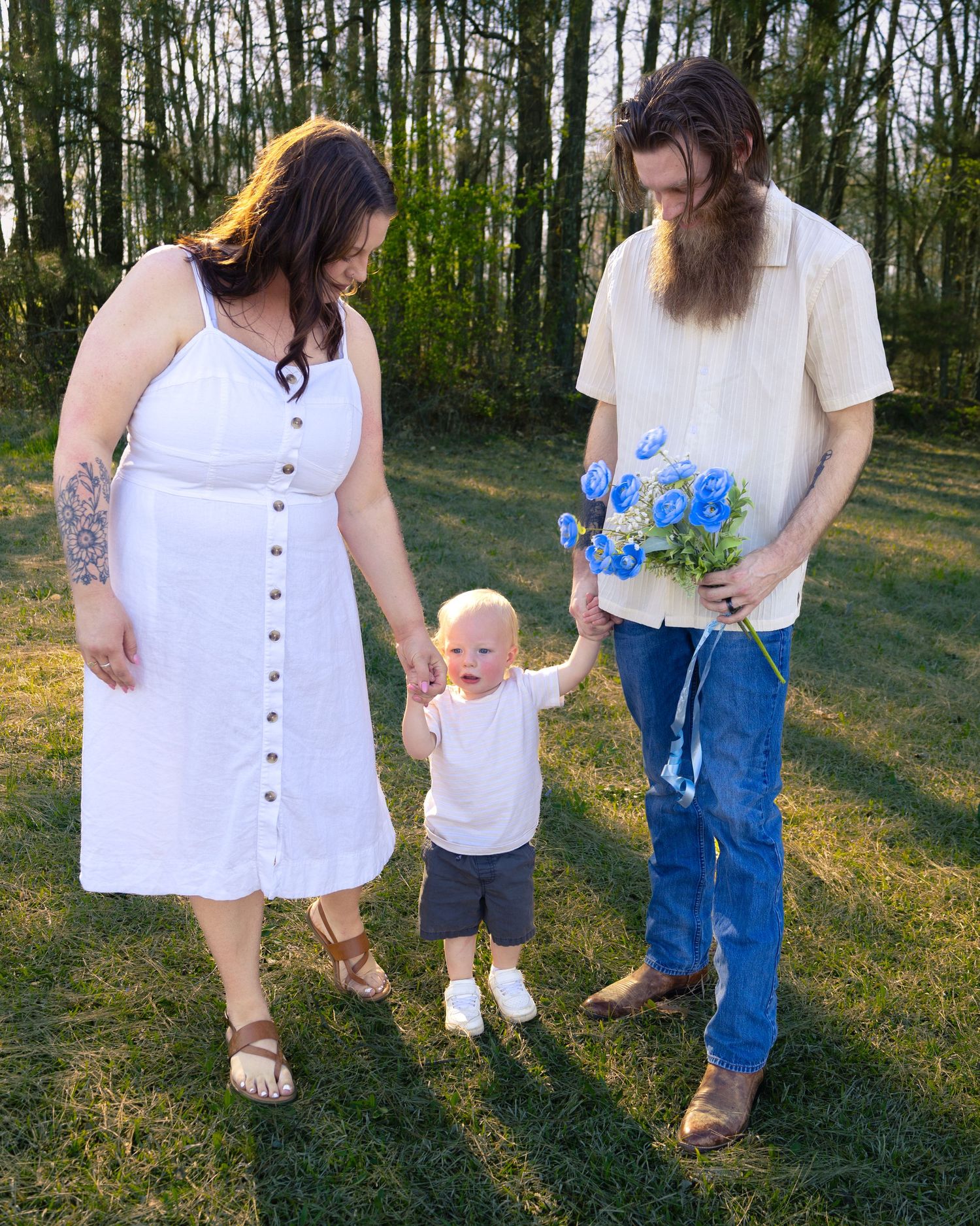 A young family with a bearded man holding blue flowers, a woman in a white dress, and a toddler standing on green grass.