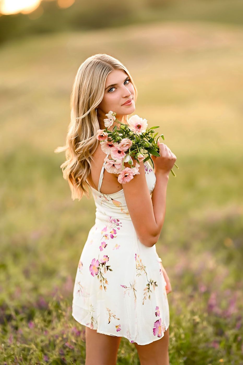 Young blonde woman in floral dress holding pink flowers in a sunlit meadow field at golden hour.
