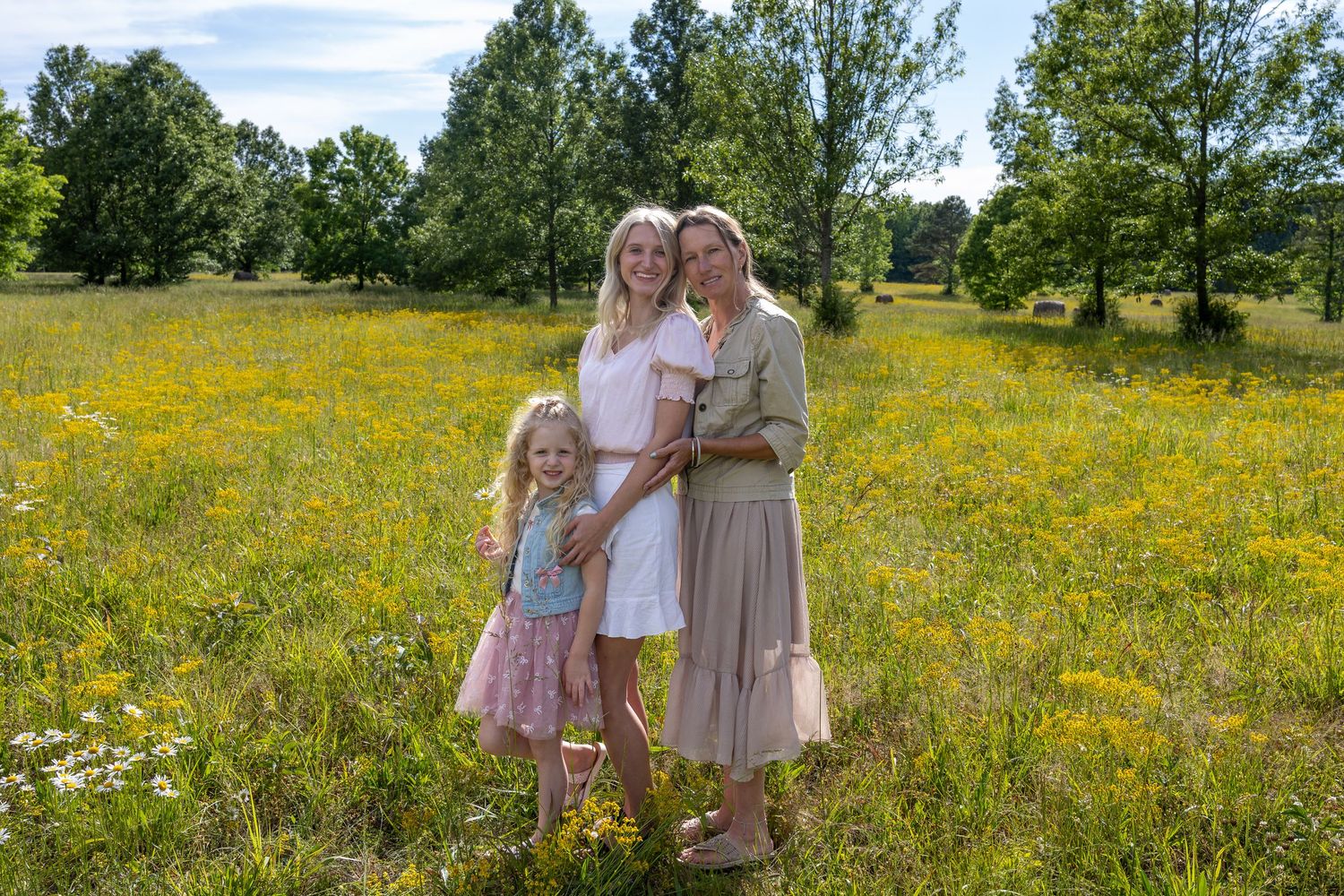 Three females pose in a sunny wildflower meadow with yellow blooms and green trees in the background.