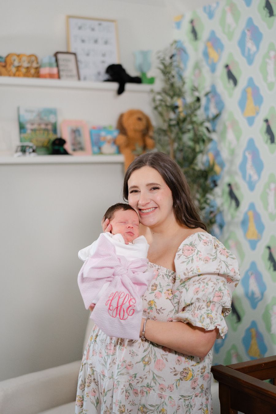 Smiling mother holding newborn baby with monogrammed pink bow in floral nursery with dog-themed wallpaper.