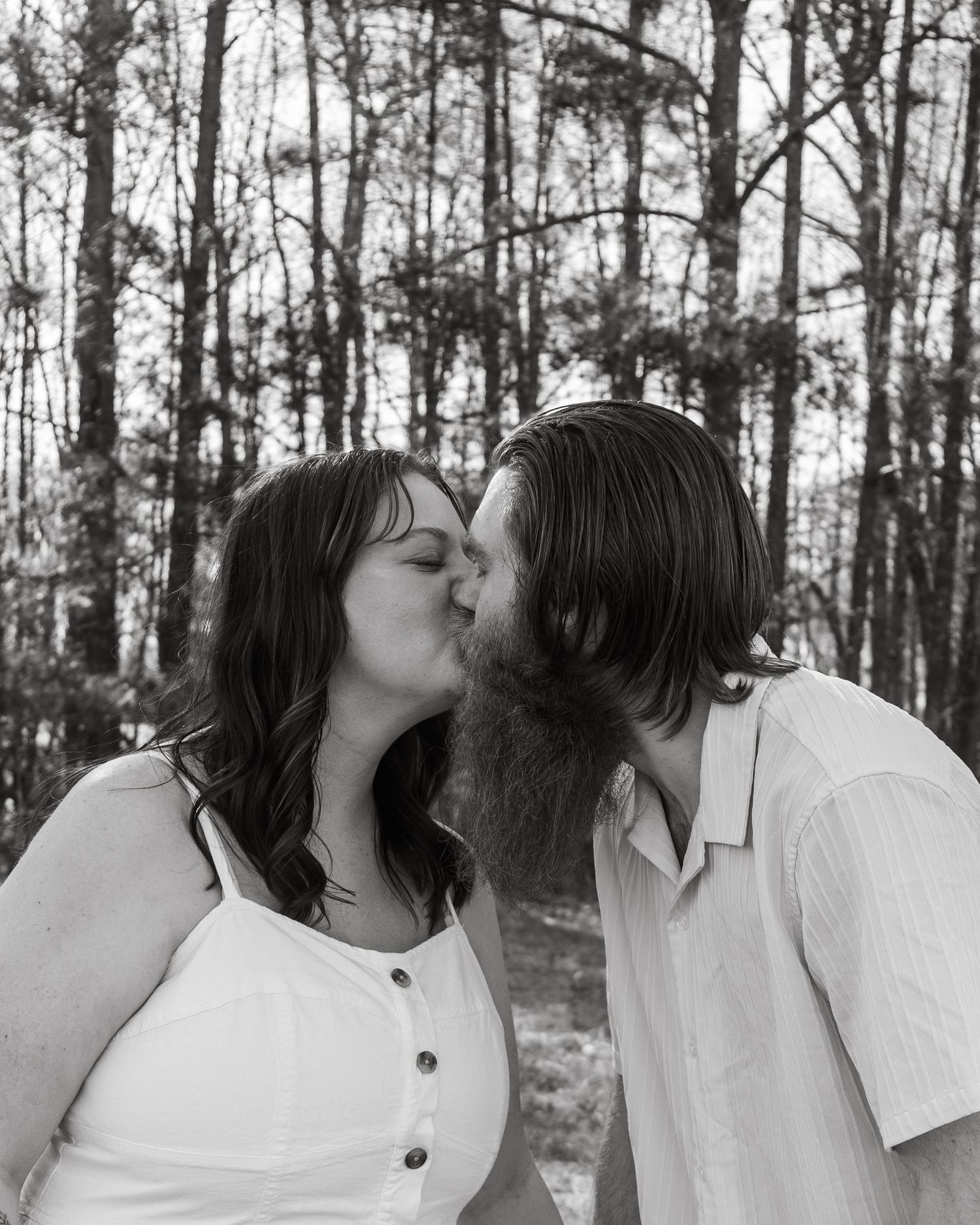 Black and white photo of a couple sharing a romantic kiss outdoors among tall trees in a forest setting.