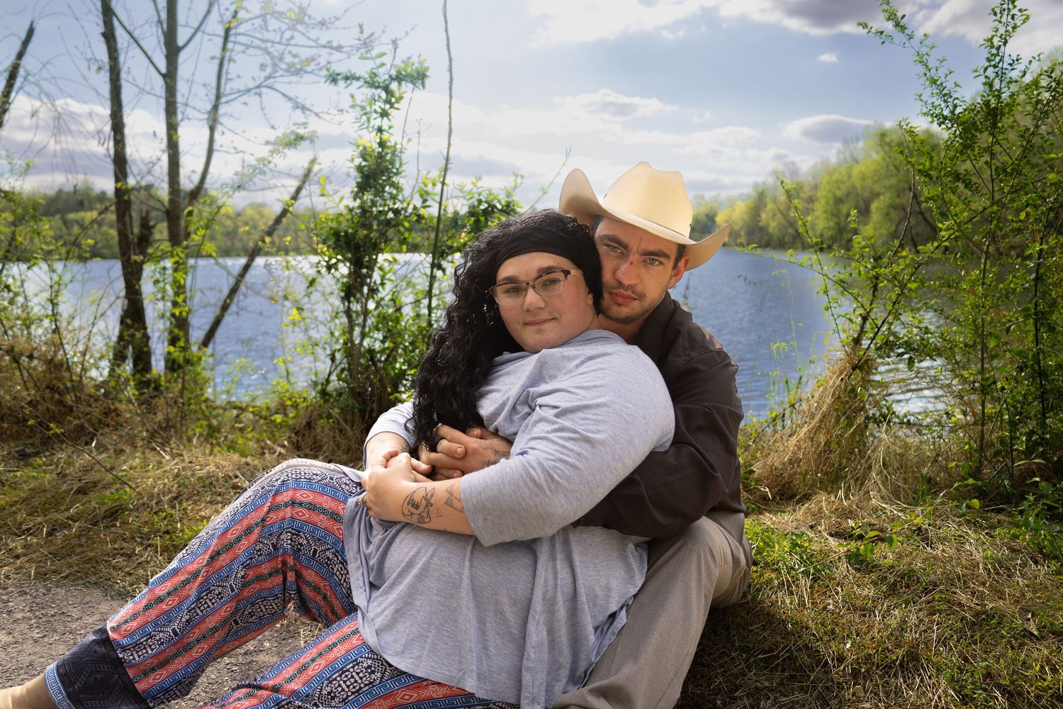 Couple embracing outdoors by a serene lake, woman in colorful pants and glasses, man in straw hat, surrounded by trees.