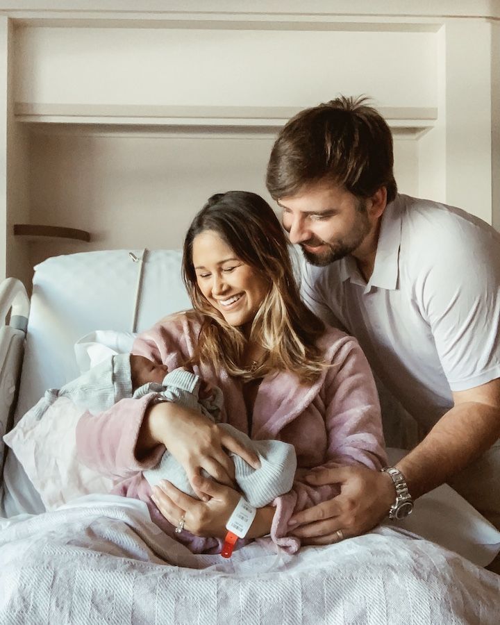 New parents smile joyfully at their newborn baby in a hospital room, mother in pink robe cradled in bed.