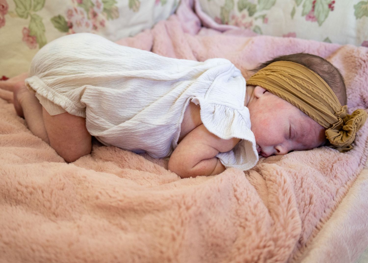 Newborn baby sleeping peacefully on a pink fluffy blanket, wearing a white outfit and mustard yellow headband bow.