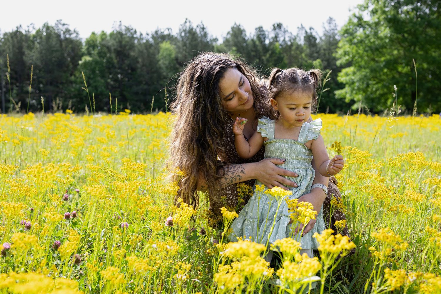 A mother and young daughter enjoy yellow wildflowers in a sunny meadow surrounded by green trees.