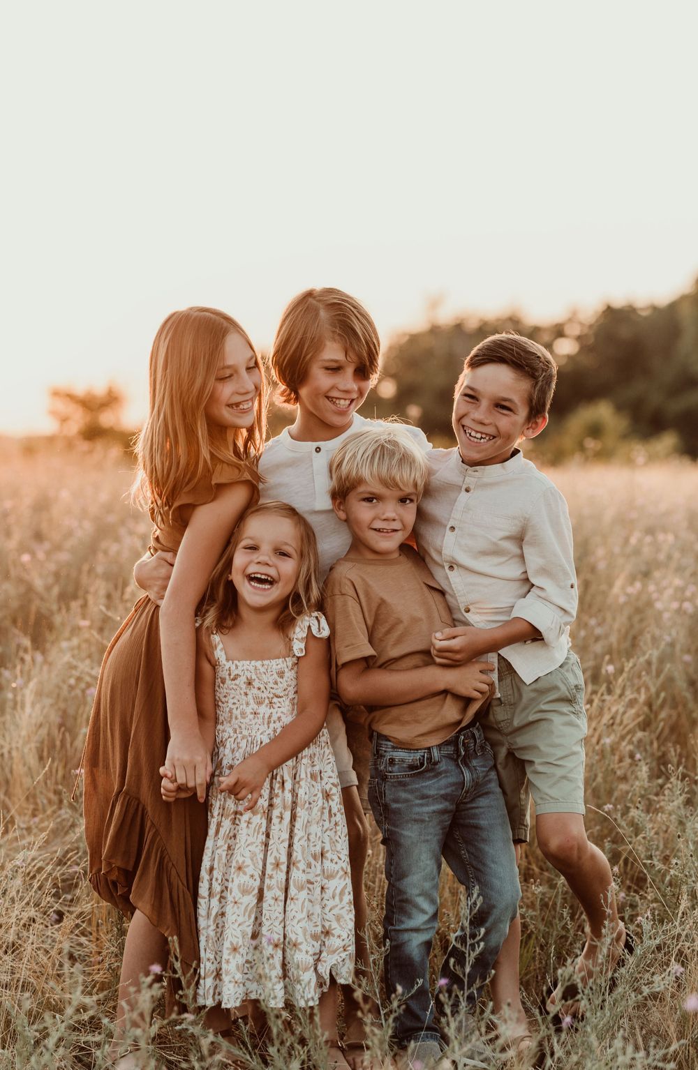 Five happy children posing together in a golden field at sunset, smiling and laughing in warm-toned outfits.