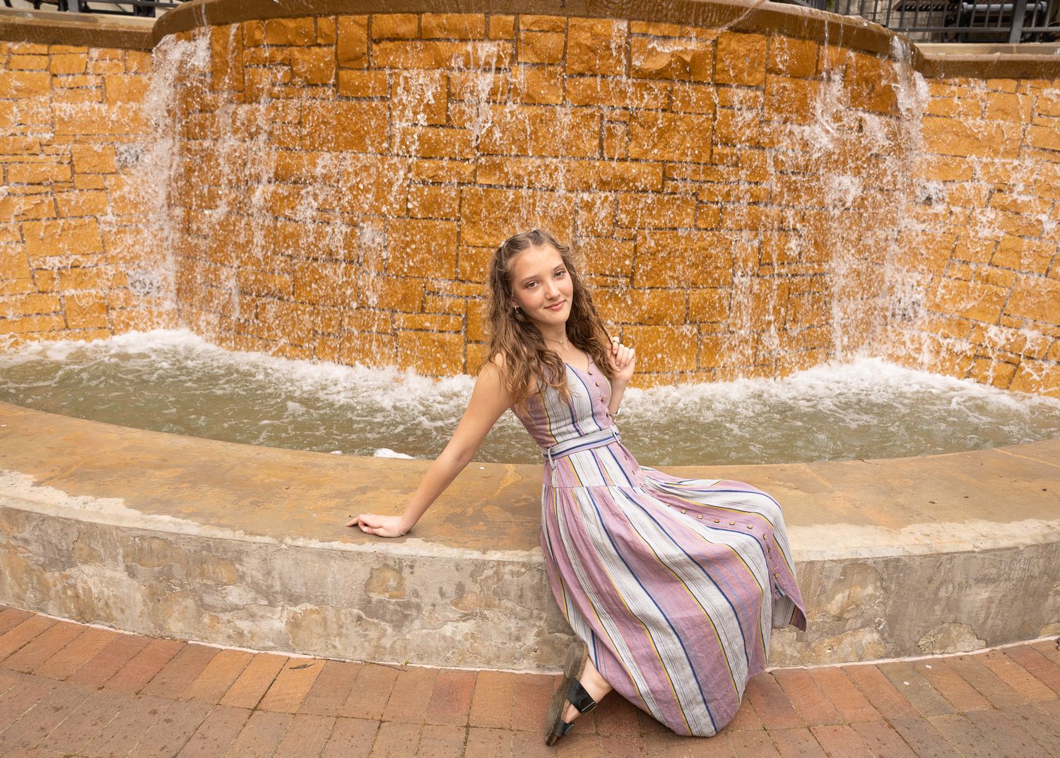 Young woman in striped maxi dress posing by a stone waterfall fountain on a brick plaza.