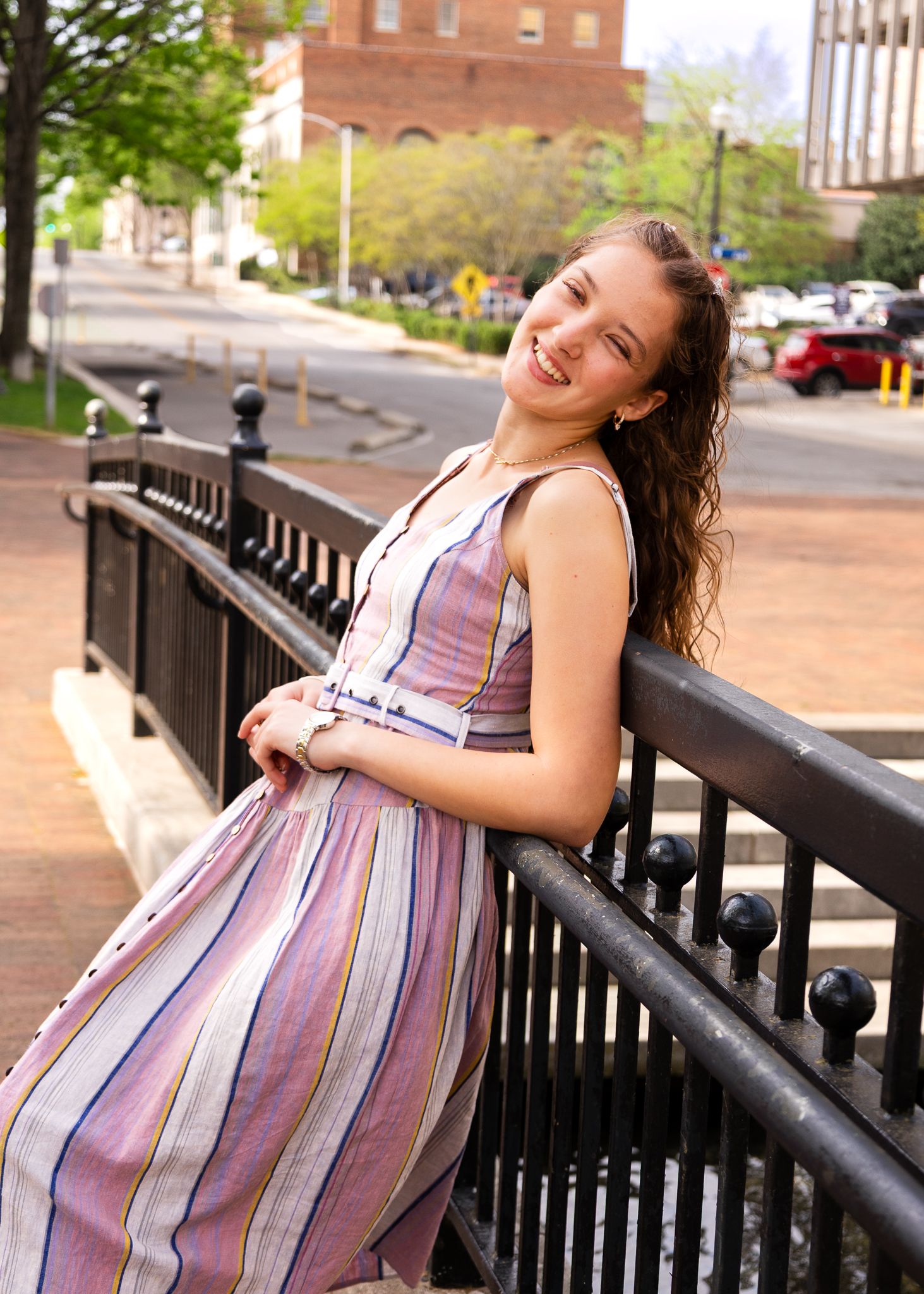 Smiling young woman in striped sundress leaning on black iron railing on urban street with trees and brick buildings.