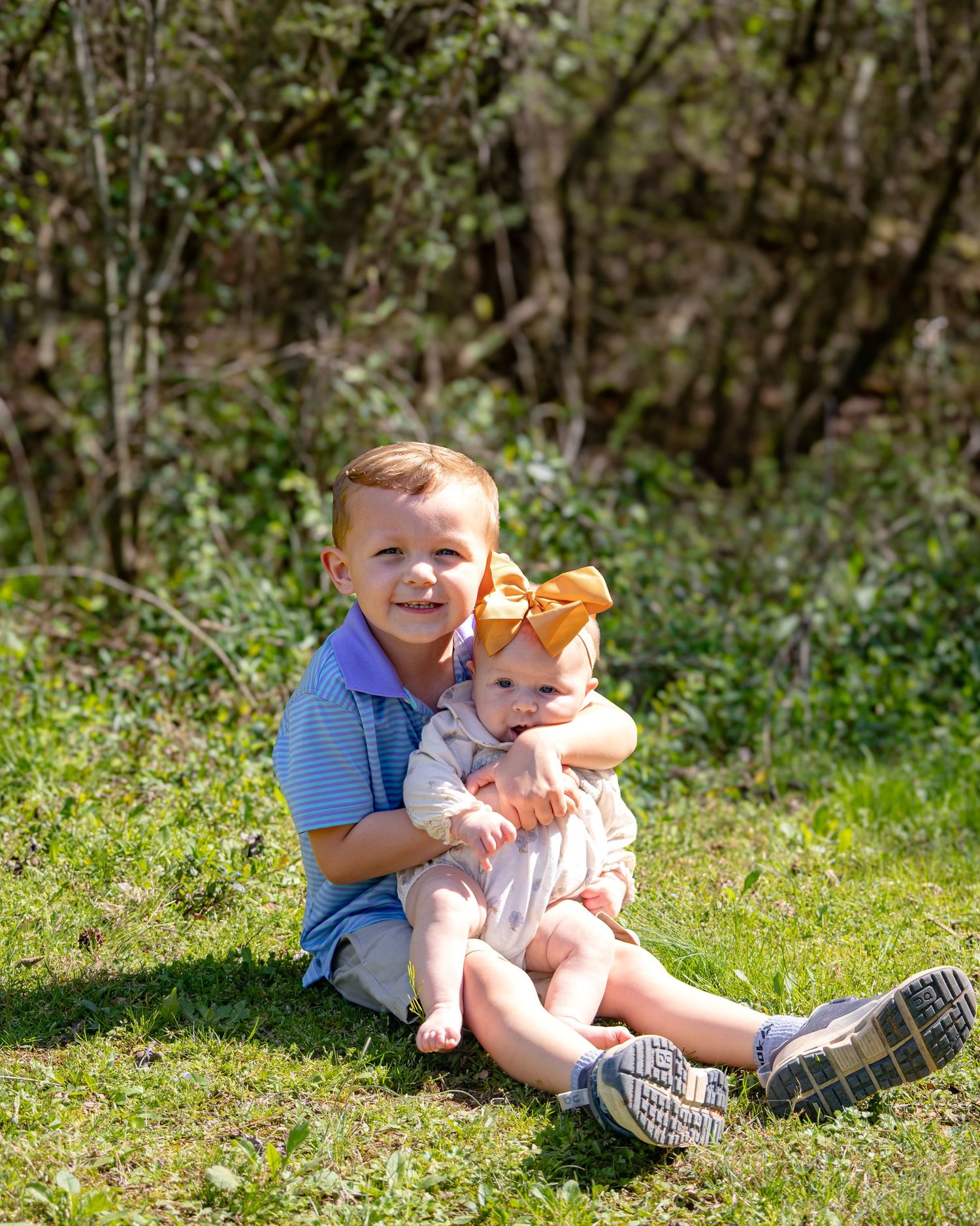 Two young children sit together on grass in a sunny outdoor setting during springtime.