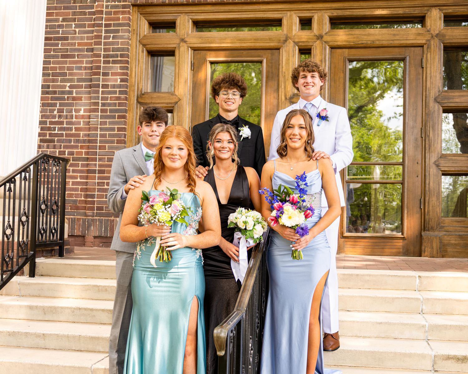 Six prom-dressed teens pose on brick building steps, girls in blue gowns with bouquets, boys in suits.