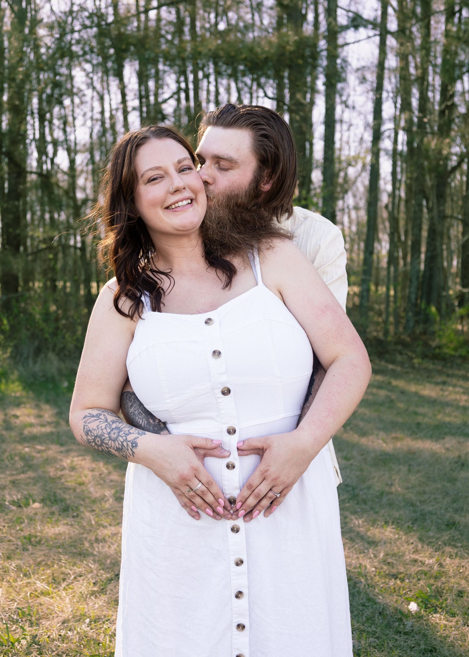 Couple posing outdoors, woman in white button dress with tattoo, man kissing her cheek, hands on baby bump.