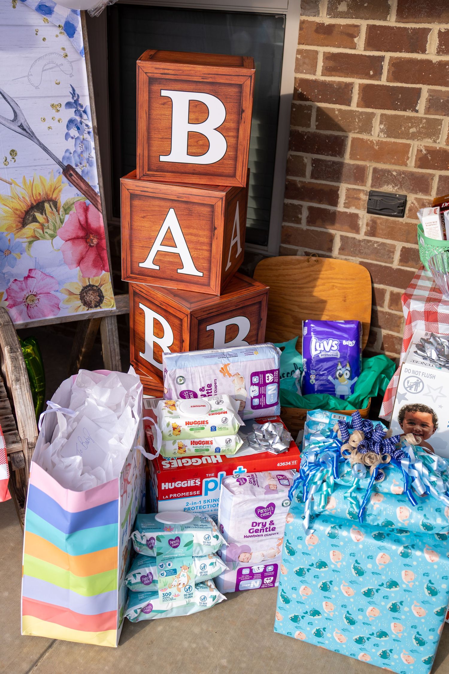 Baby shower display with wooden letter blocks spelling BABY, Huggies wipes, Luvs diapers, and colorful gift bags on brick steps.
