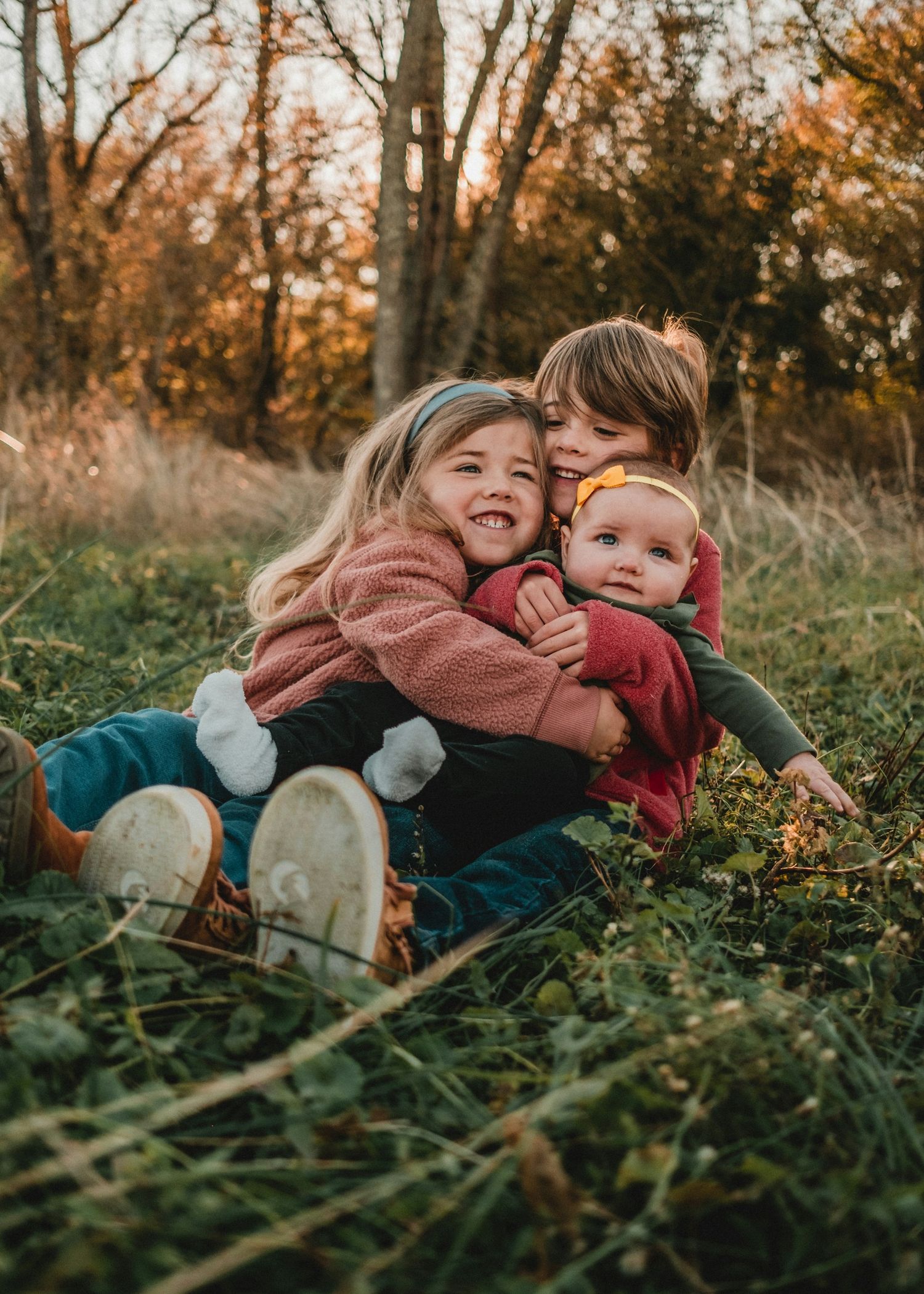 Three siblings hugging and smiling together while sitting in a grassy field during golden autumn sunset.