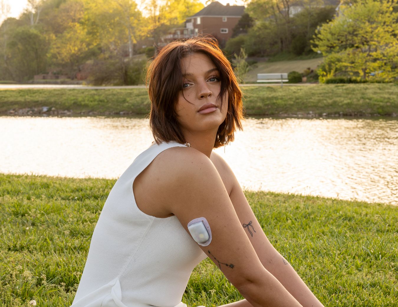 Woman sitting on grass by a lake at sunset, wearing a white tank top with a medical patch on her arm.