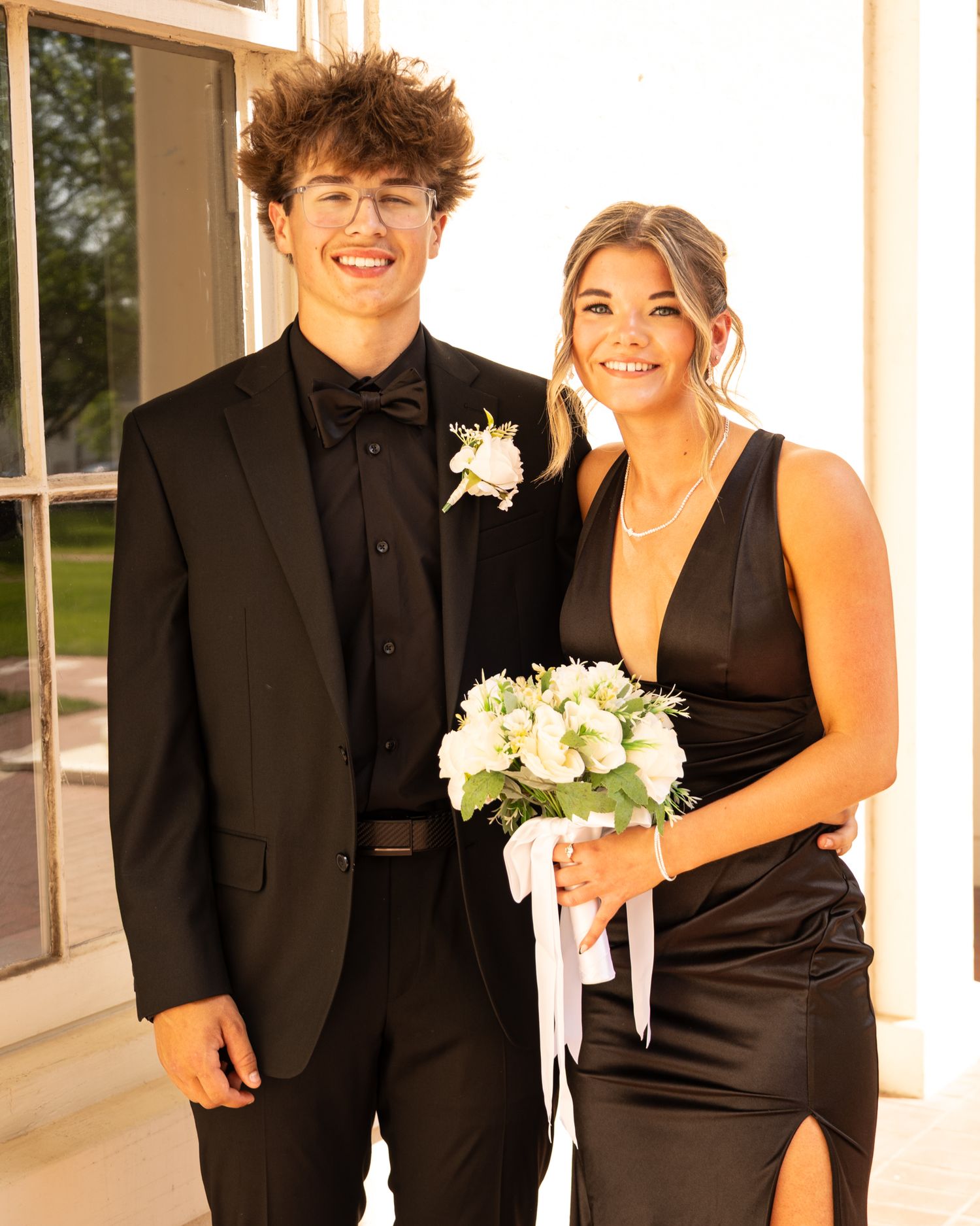 Young couple in matching black formal wear posing together, man in suit with boutonniere, woman holding white bouquet.