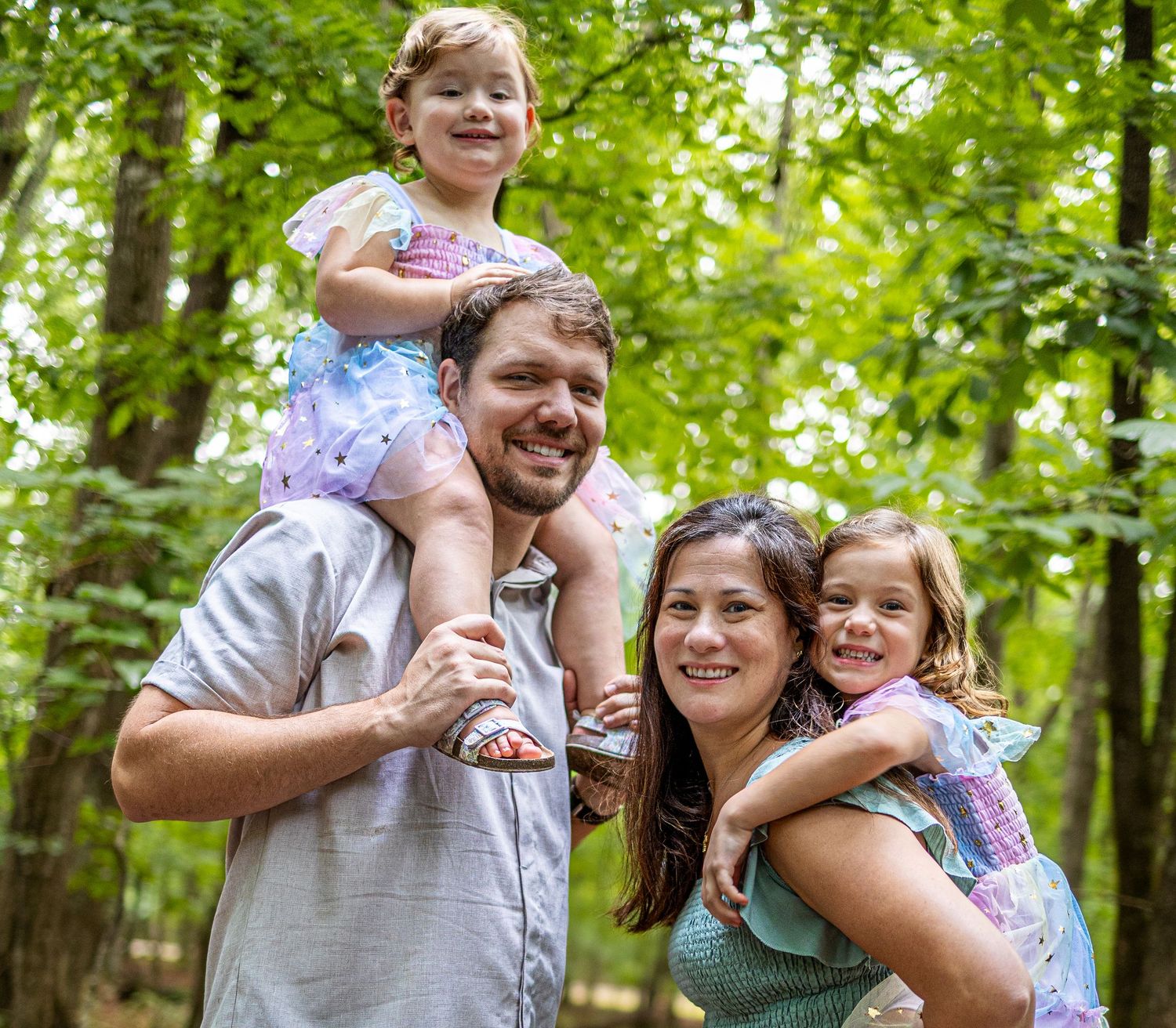 A smiling family of four poses outdoors in a lush green forest, with two young girls riding on their parents shoulders.