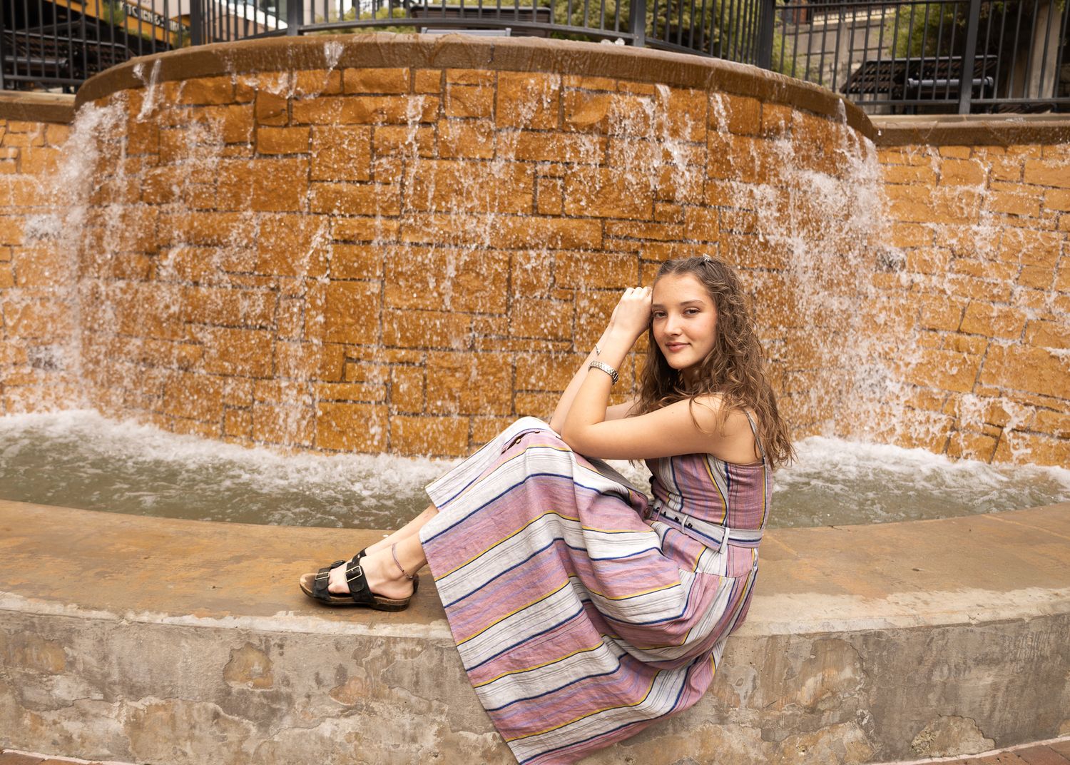 Young woman in striped maxi dress poses gracefully by a stone waterfall fountain outdoors.
