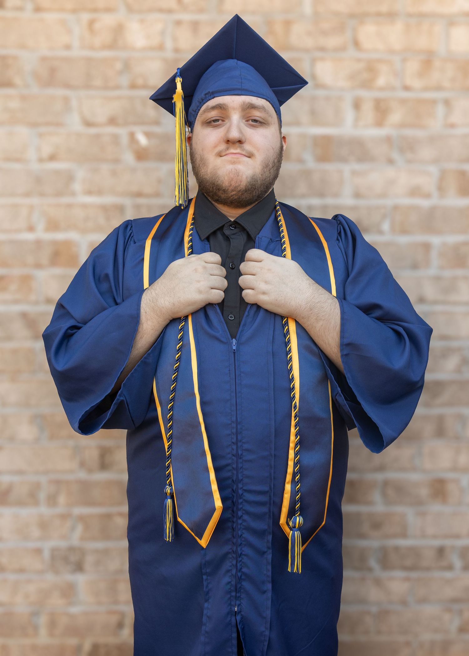 Young man in navy blue graduation cap and gown with gold honor cords poses proudly in front of brick wall.
