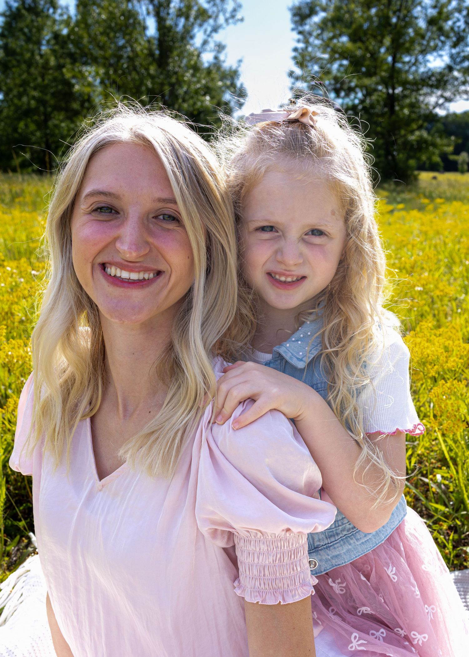 A smiling blonde woman and young girl with curly hair pose together in a sunny yellow wildflower field.
