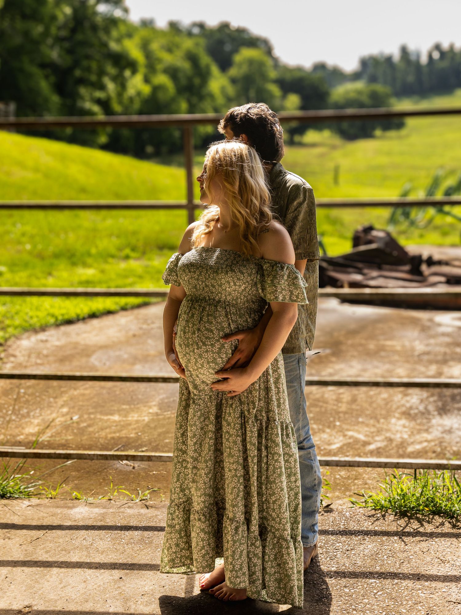 Couple sharing a tender maternity moment outdoors on a farm, woman in floral dress cradling her baby bump.