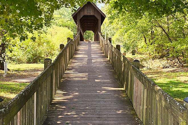 Wooden covered bridge with railings leads through lush green trees on a sunny day.