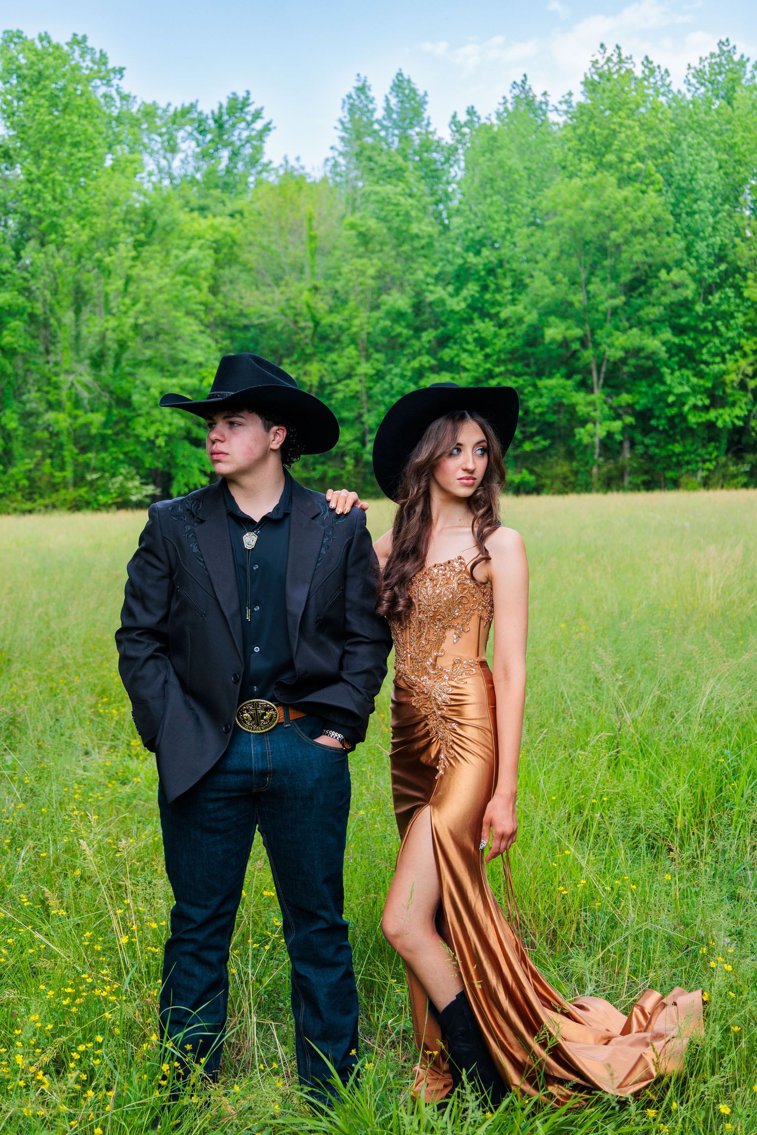 A young couple in black cowboy hats pose in a green field, he in a suit, she in a gold gown.
