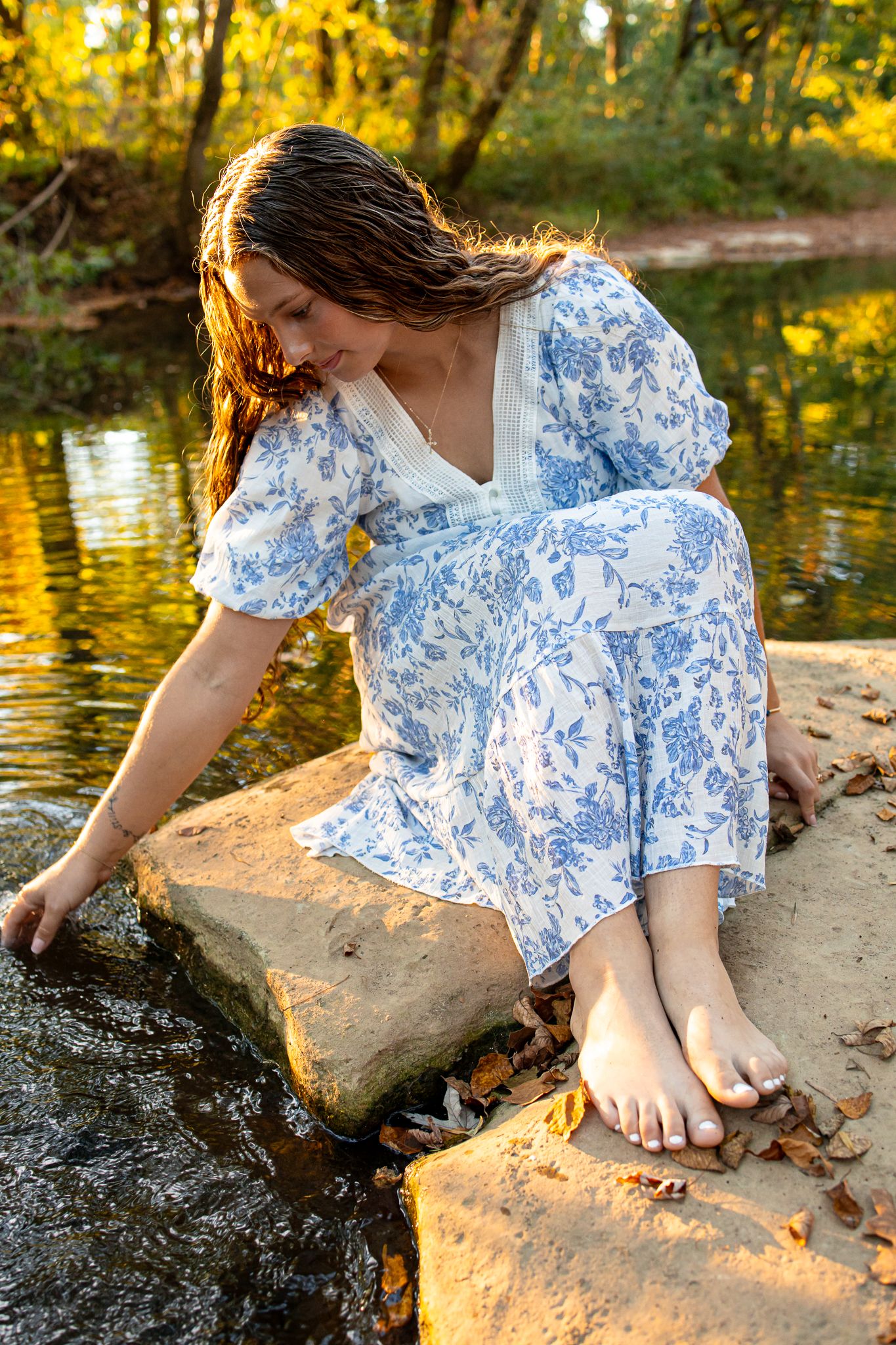 Woman in blue floral dress sitting barefoot on rocks by a serene creek surrounded by autumn foliage.