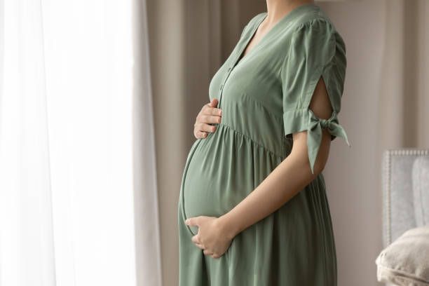 Pregnant woman in sage green dress gently cradling her baby bump while standing near a window during her maternity shoot in Madison Alabama