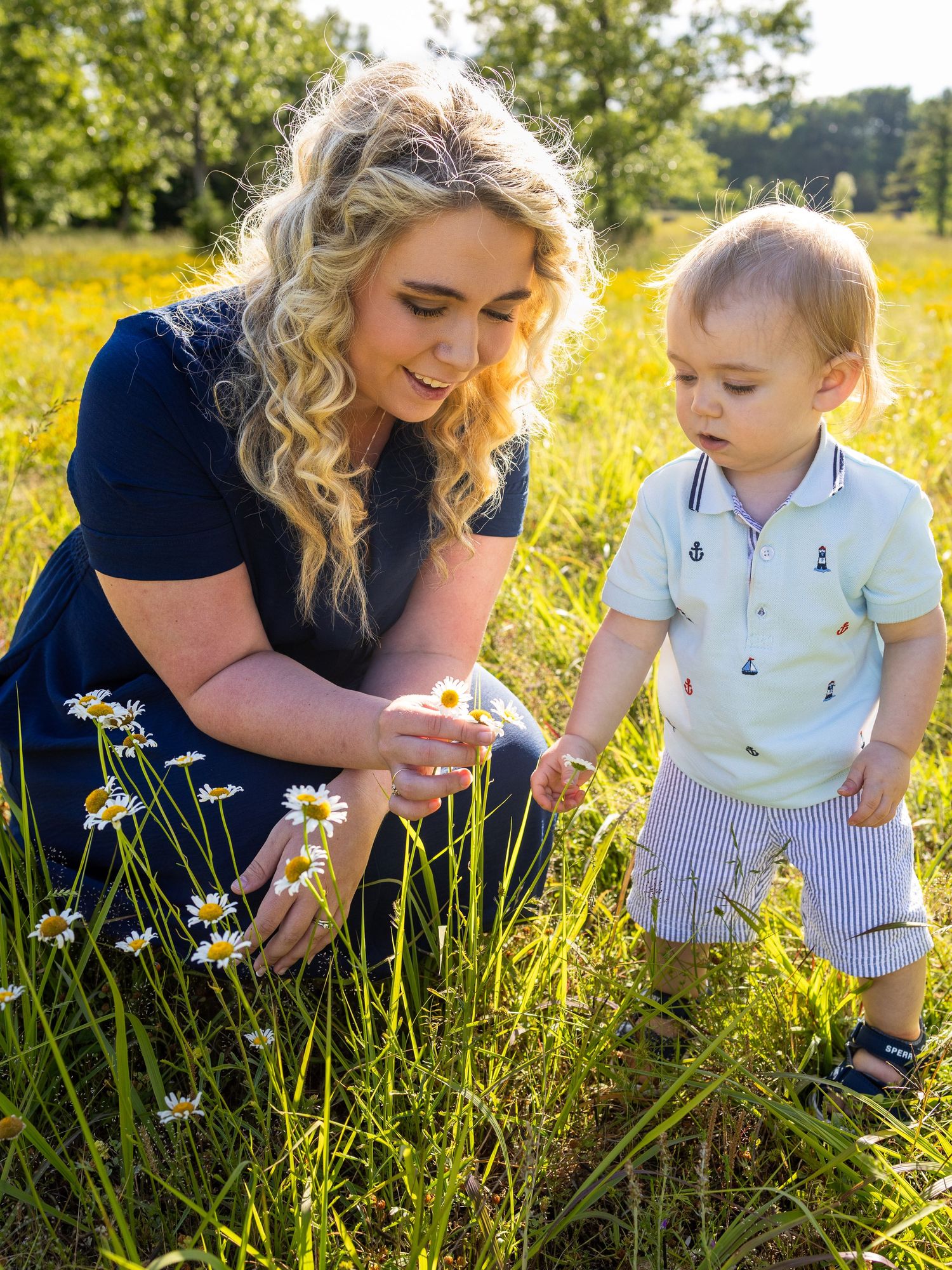 A woman with curly blonde hair and a toddler boy examine white daisies together in a sunny meadow.