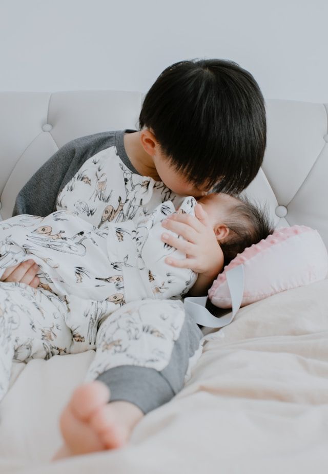 An older child in pajamas gently kisses a sleeping newborn baby resting on a pink pillow on a white bed.