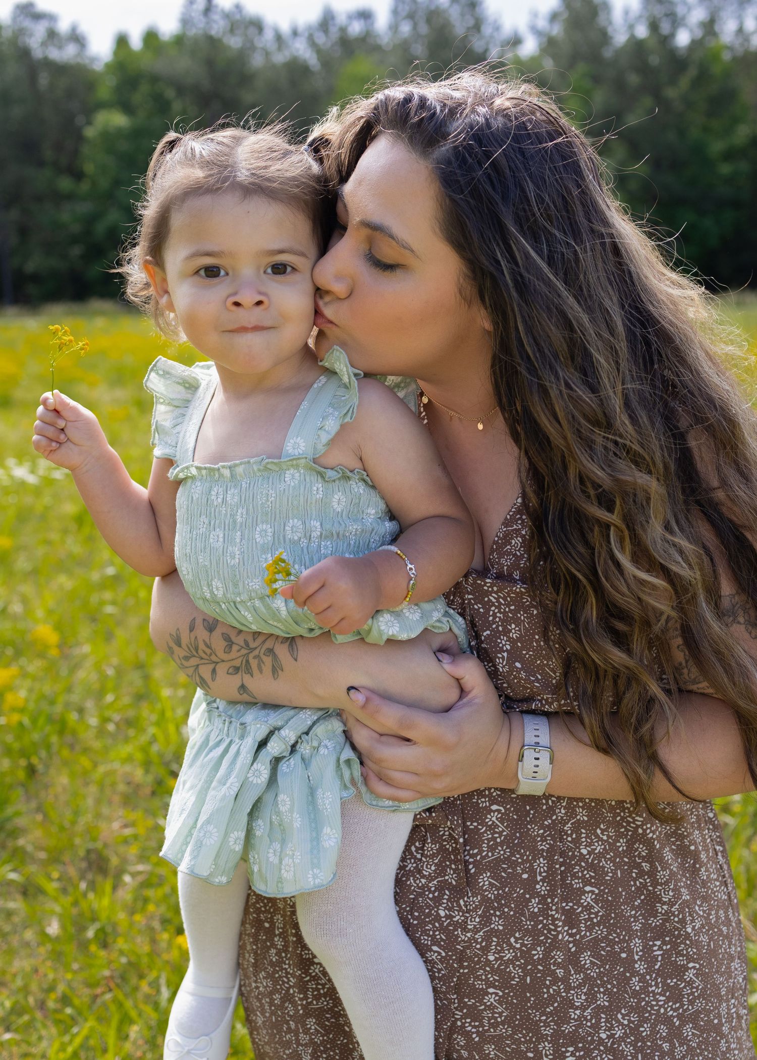 A mother kisses her toddler daughter on the cheek while holding her in a sunny flower field.
