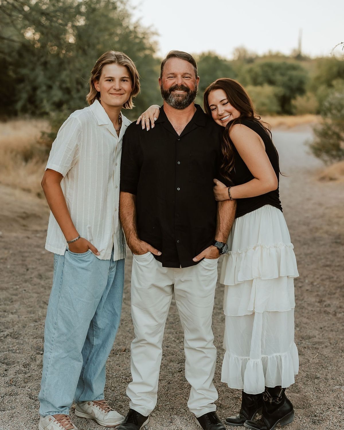A man and two teenagers smile together on a dirt path surrounded by desert greenery during golden hour.