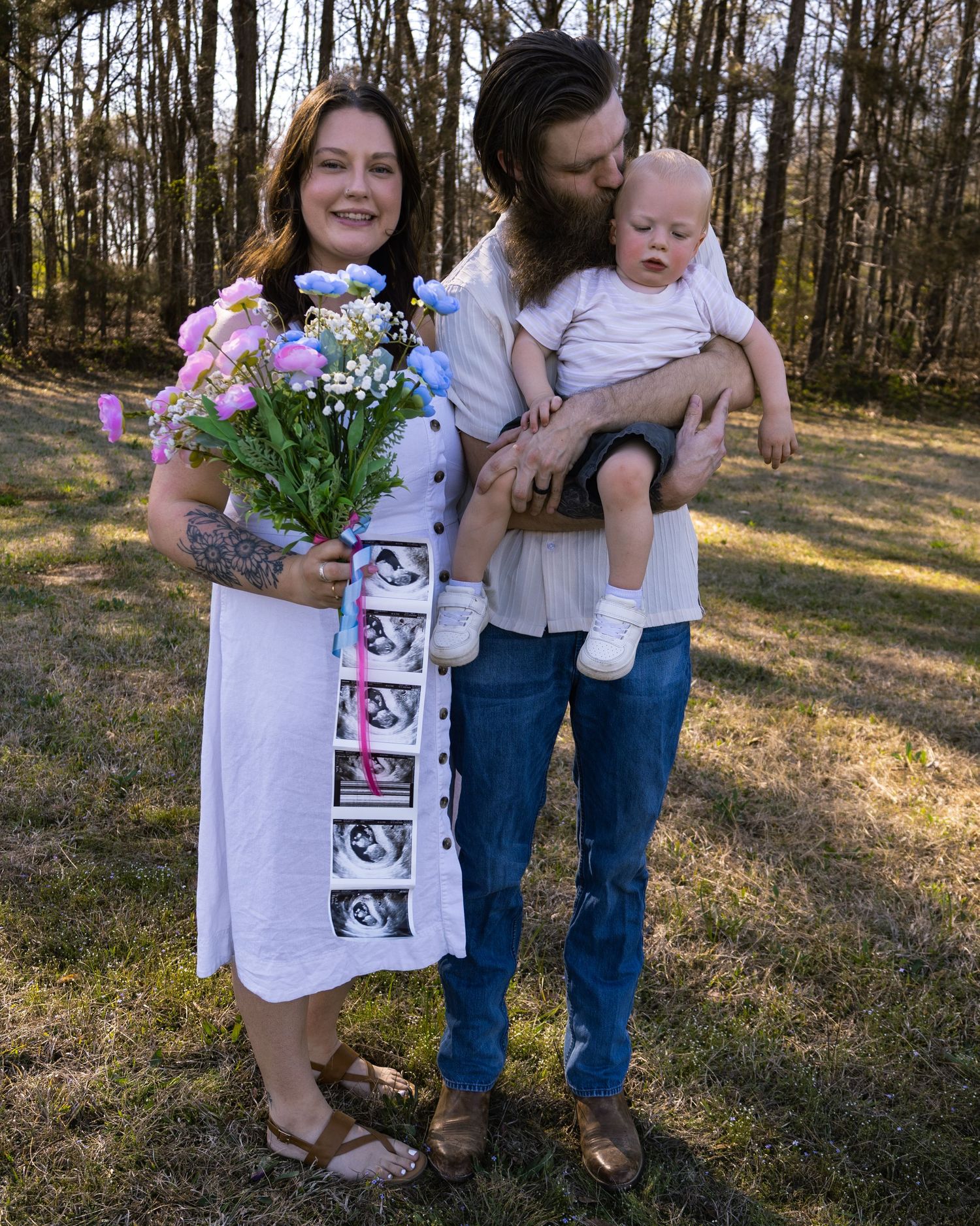Couple poses outdoors with baby and ultrasound photos for a pregnancy announcement, woman holding pink and blue flowers.