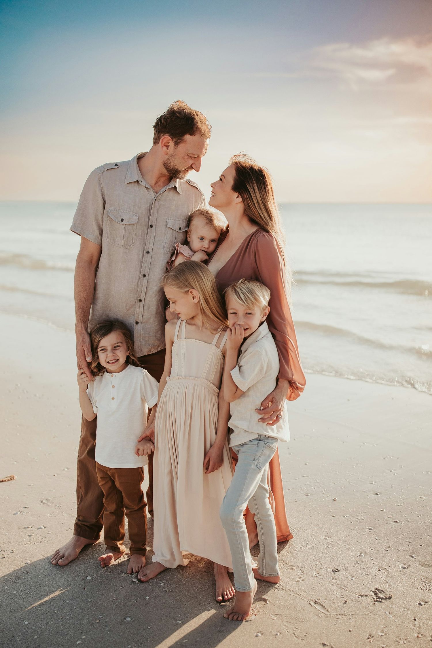 Family of six poses on a sandy beach at sunset, parents sharing a tender moment while four children smile and embrace.