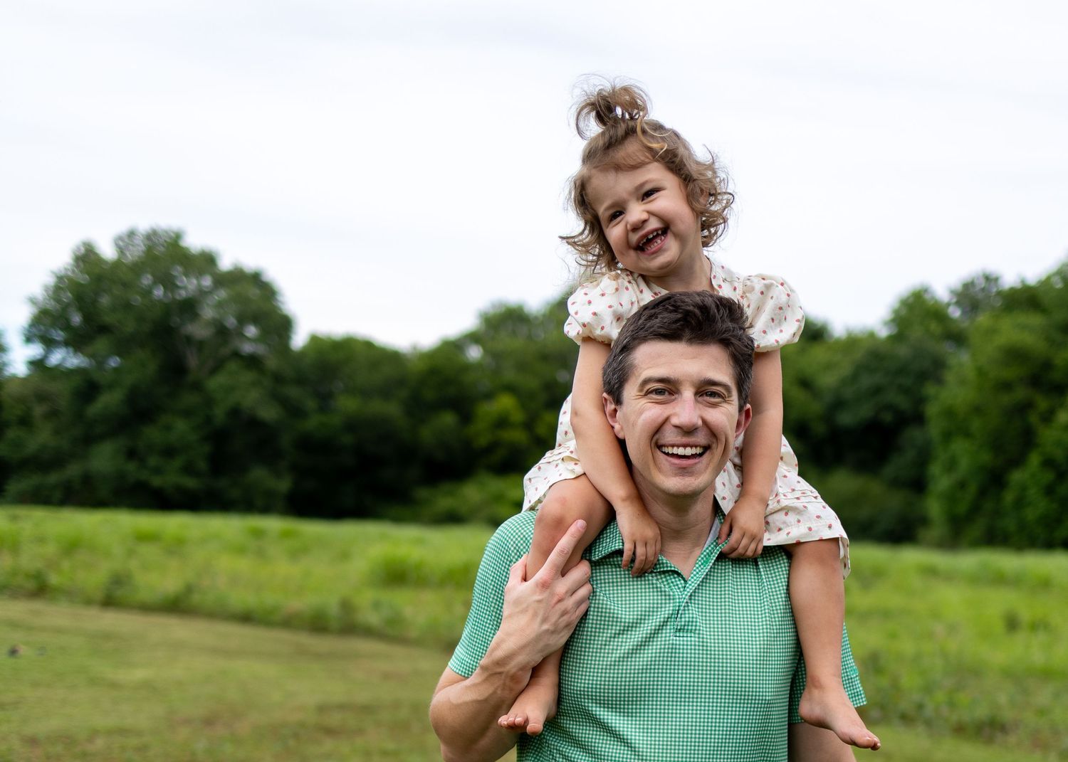 A laughing father carries his joyful young daughter on his shoulders in a lush green field outdoors.