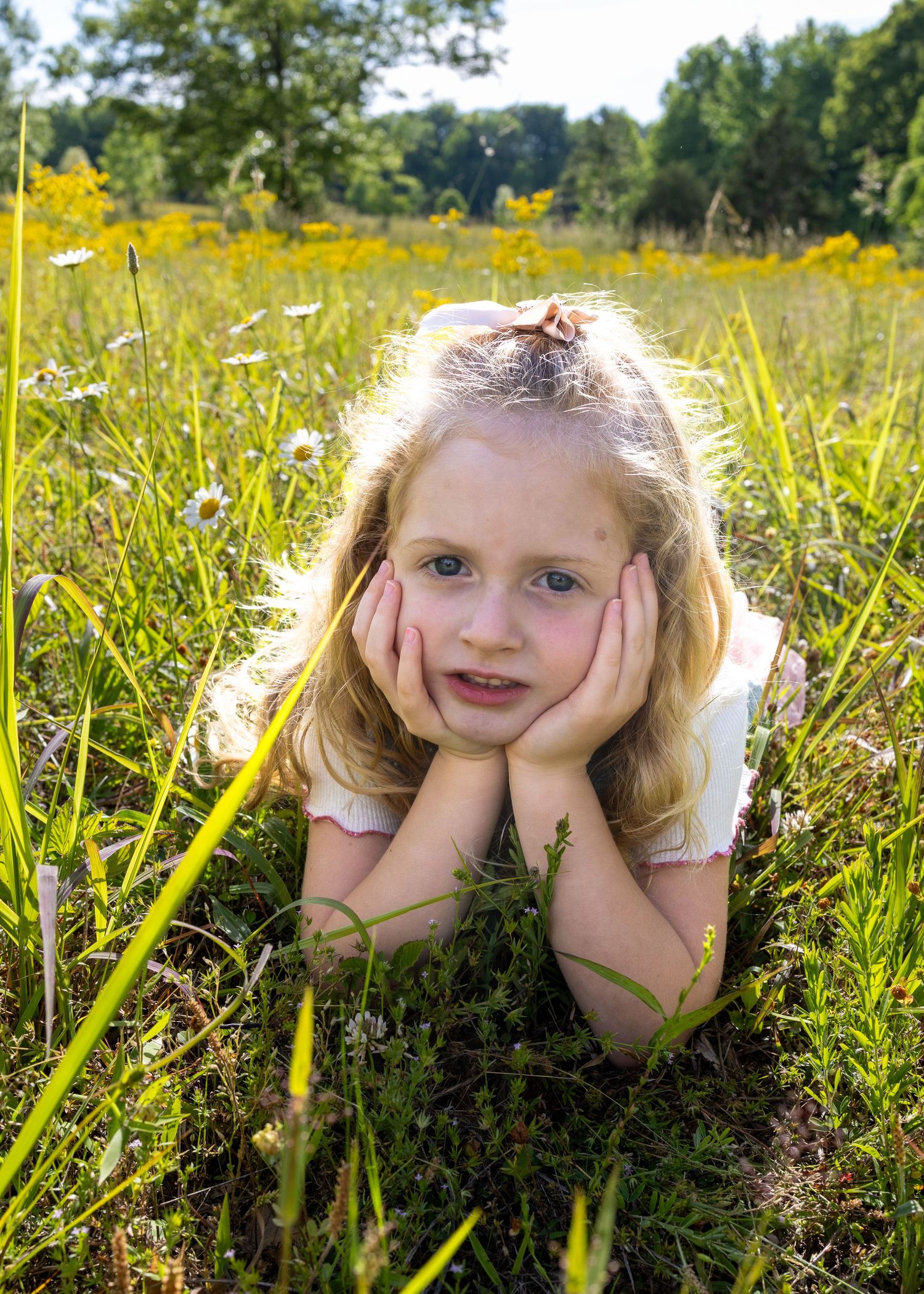 Young blonde girl lying in a sunny wildflower meadow, resting chin on hands, surrounded by green grass and yellow flowers.