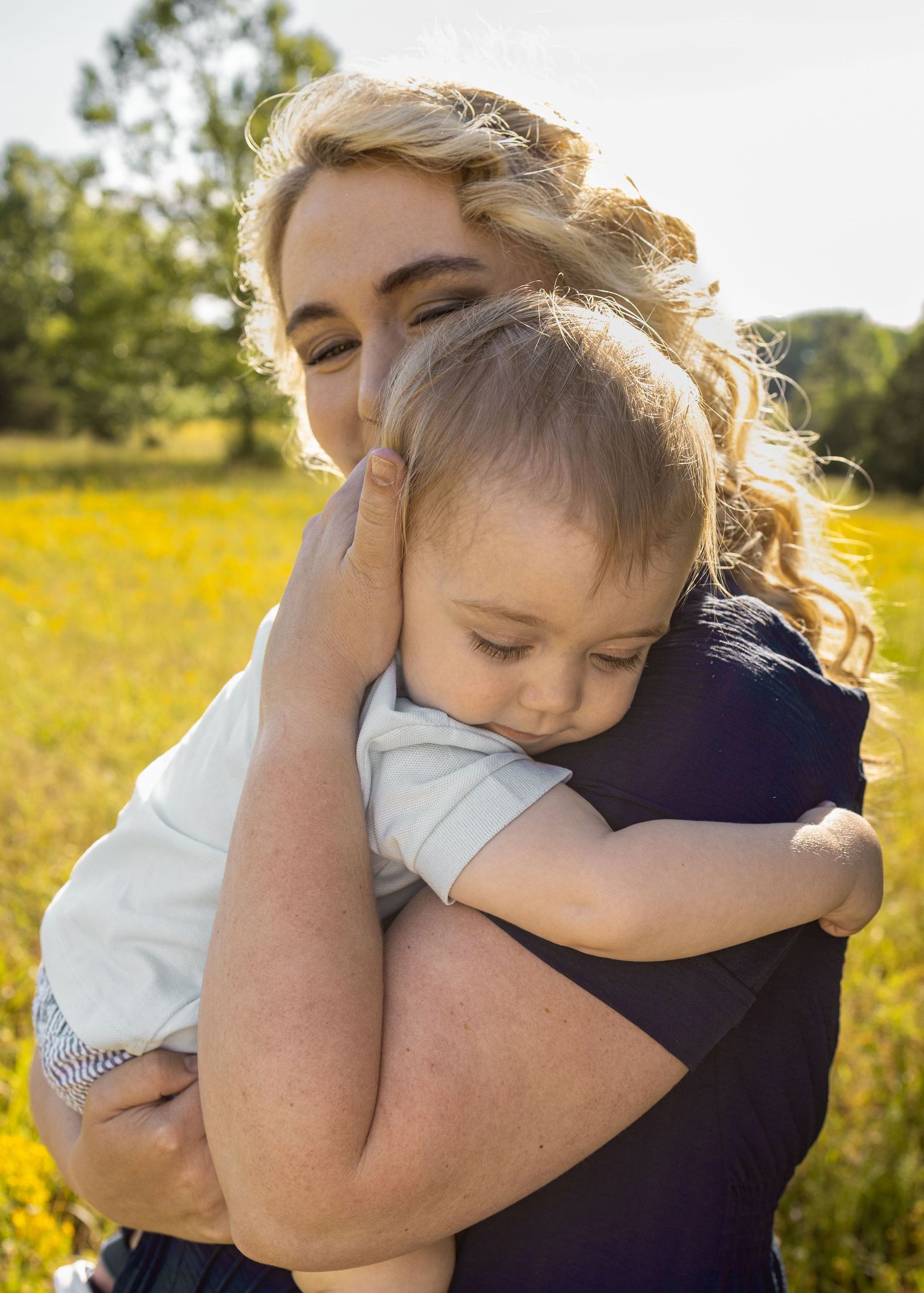 A blonde woman tenderly hugs a young toddler in a sunny field of yellow wildflowers outdoors.