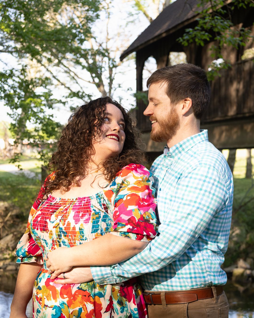 A smiling couple embraces outdoors near a covered bridge, woman in colorful floral dress, man in teal plaid shirt.
