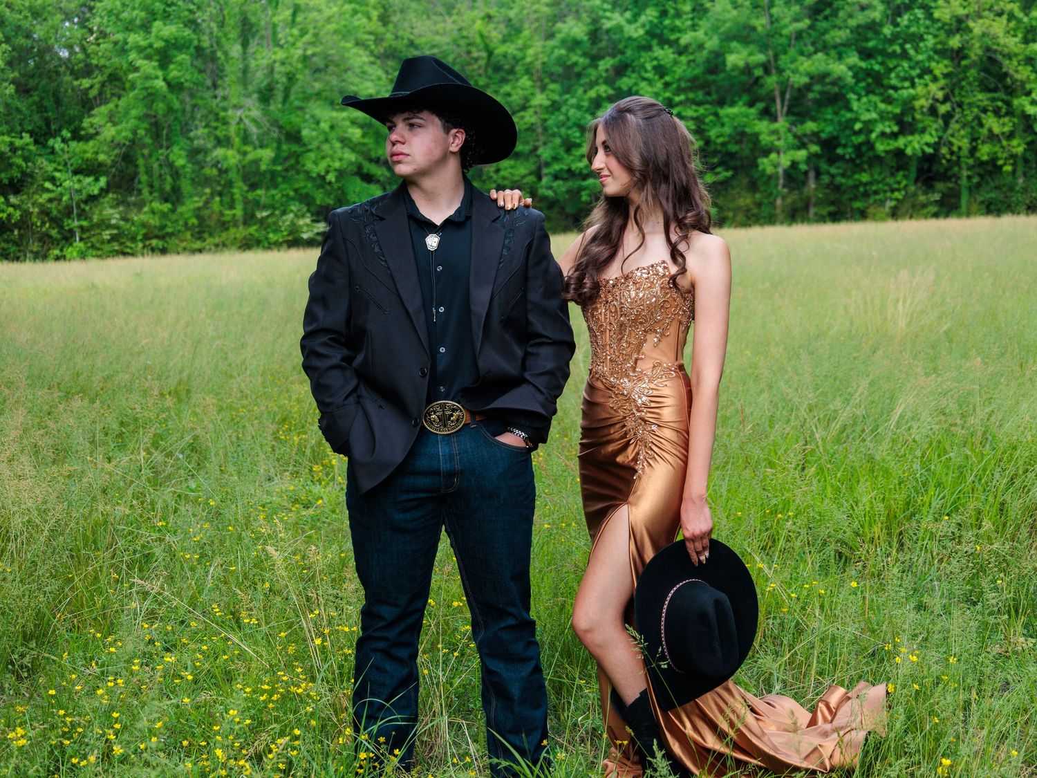 A couple in Western attire poses in a green meadow, man in black suit and cowboy hat, woman in gold sequin gown.