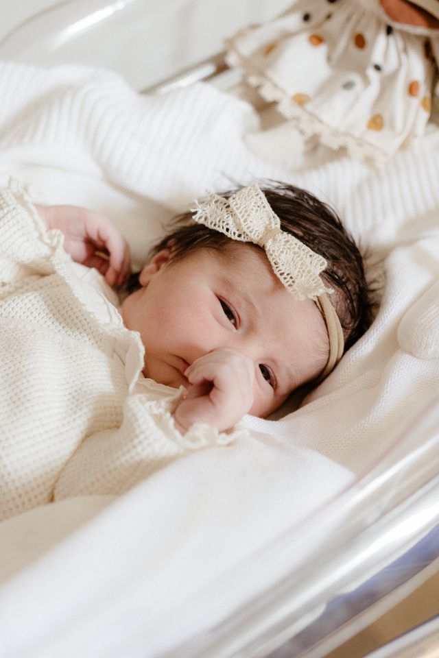Newborn baby girl in cream knit outfit and lace bow headband lying in hospital bassinet.