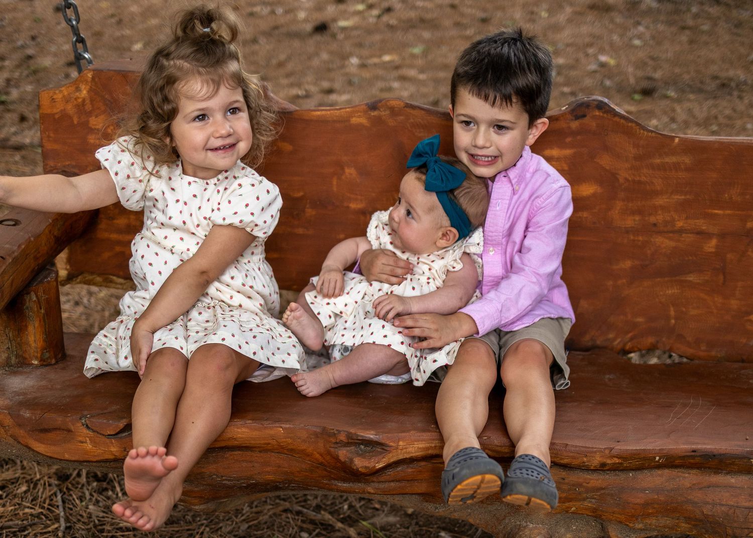 Two young children sit on a rustic wooden bench holding a newborn baby outdoors.